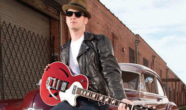 man sitting on hood of vintage car holding red Cort vintage electric guitar